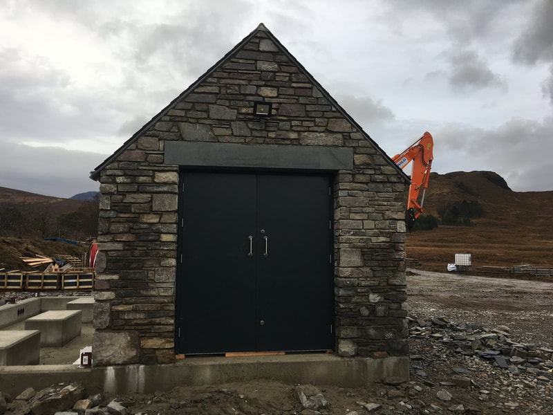 A stone Gable end with grey double doors in the middle, cloudy skies in the background